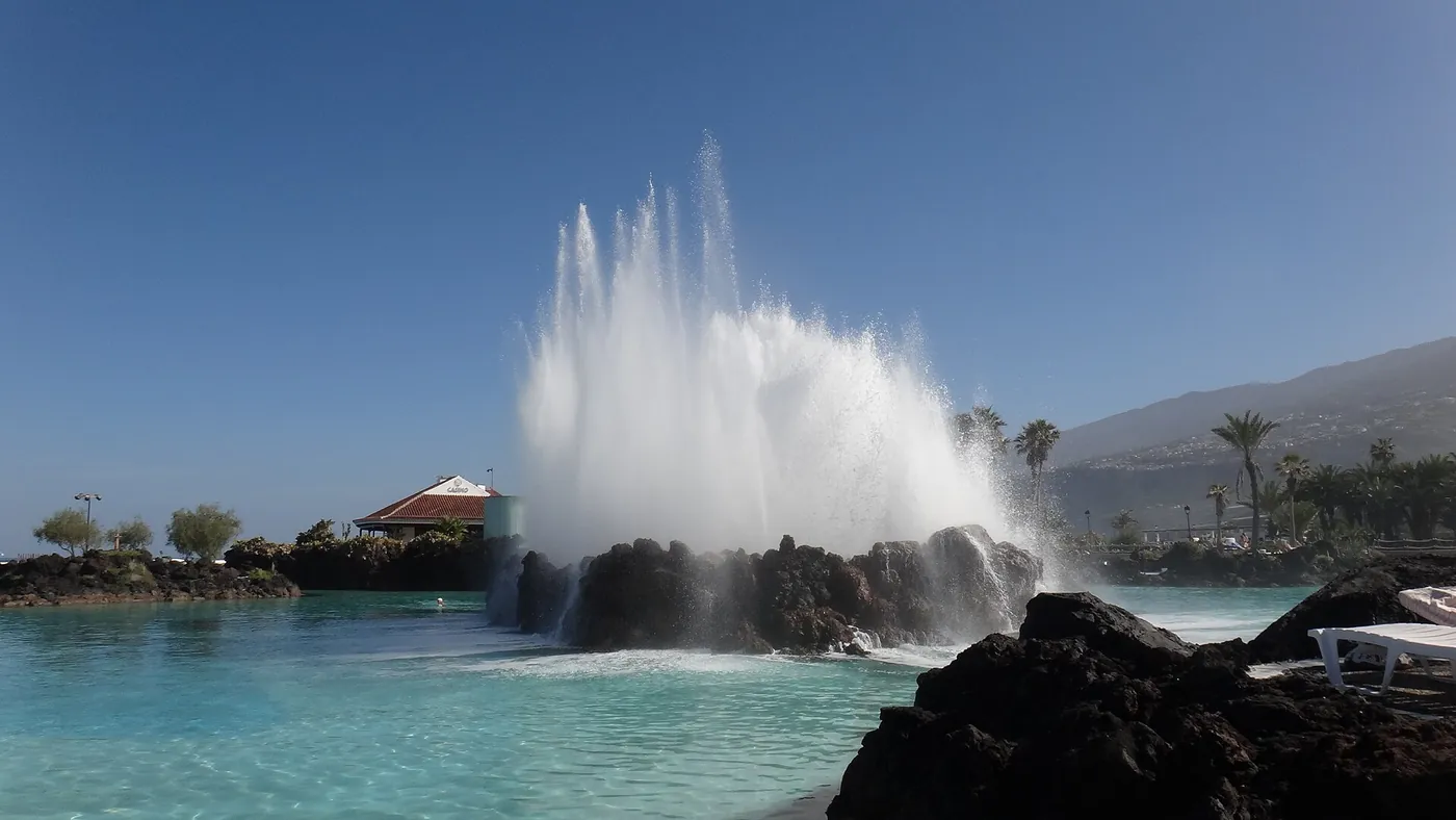 Chorro de agua de mar sobre una isla de roca volcánica en el Lago Martiánez, con las piscinas turquesa y la ladera de Puerto de la Cruz al fondo