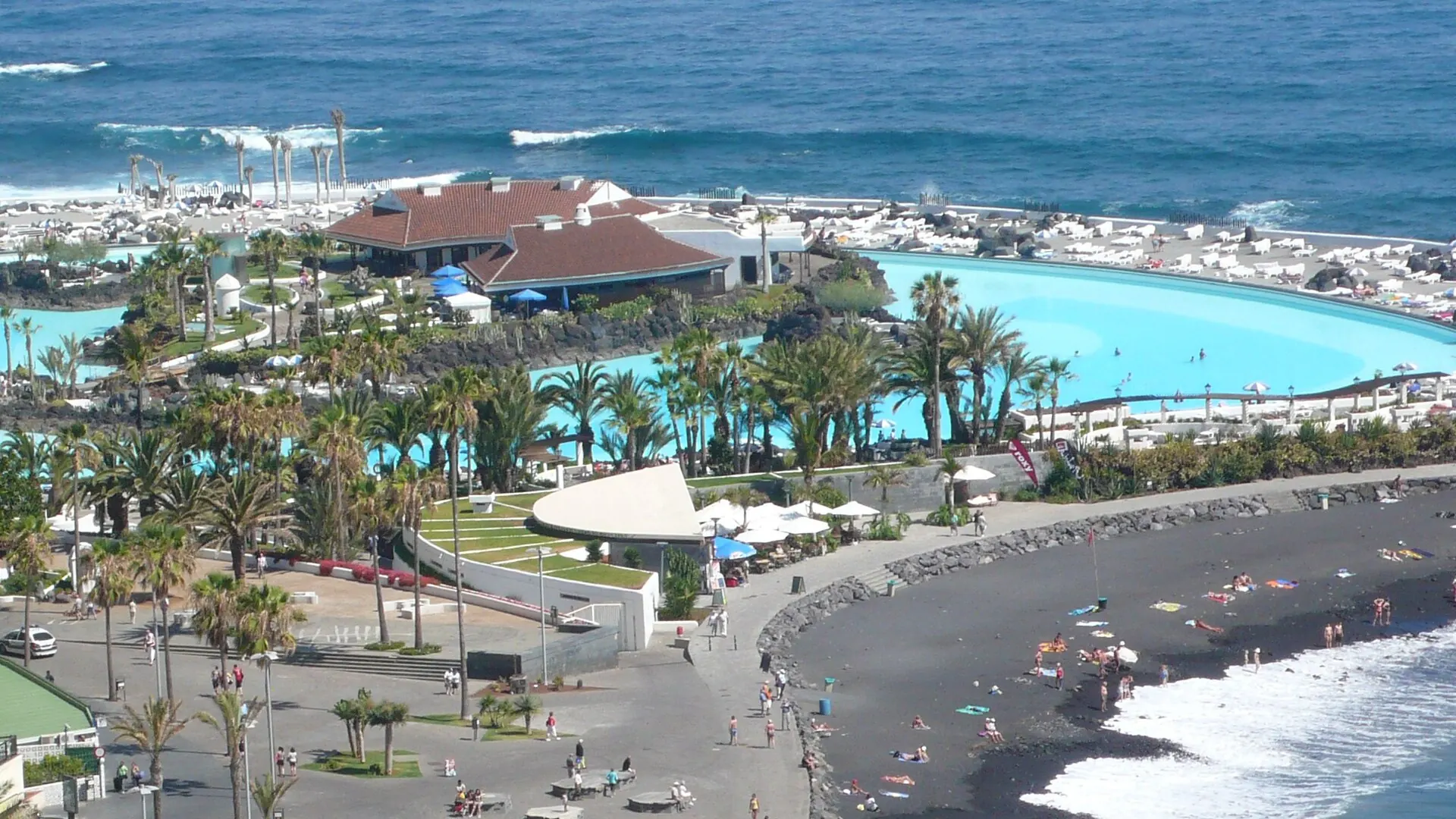 Vista aérea del Lago Martiánez con las piscinas de agua de mar, los jardines y el océano Atlántico al fondo, junto a la playa de arena negra de Martiánez