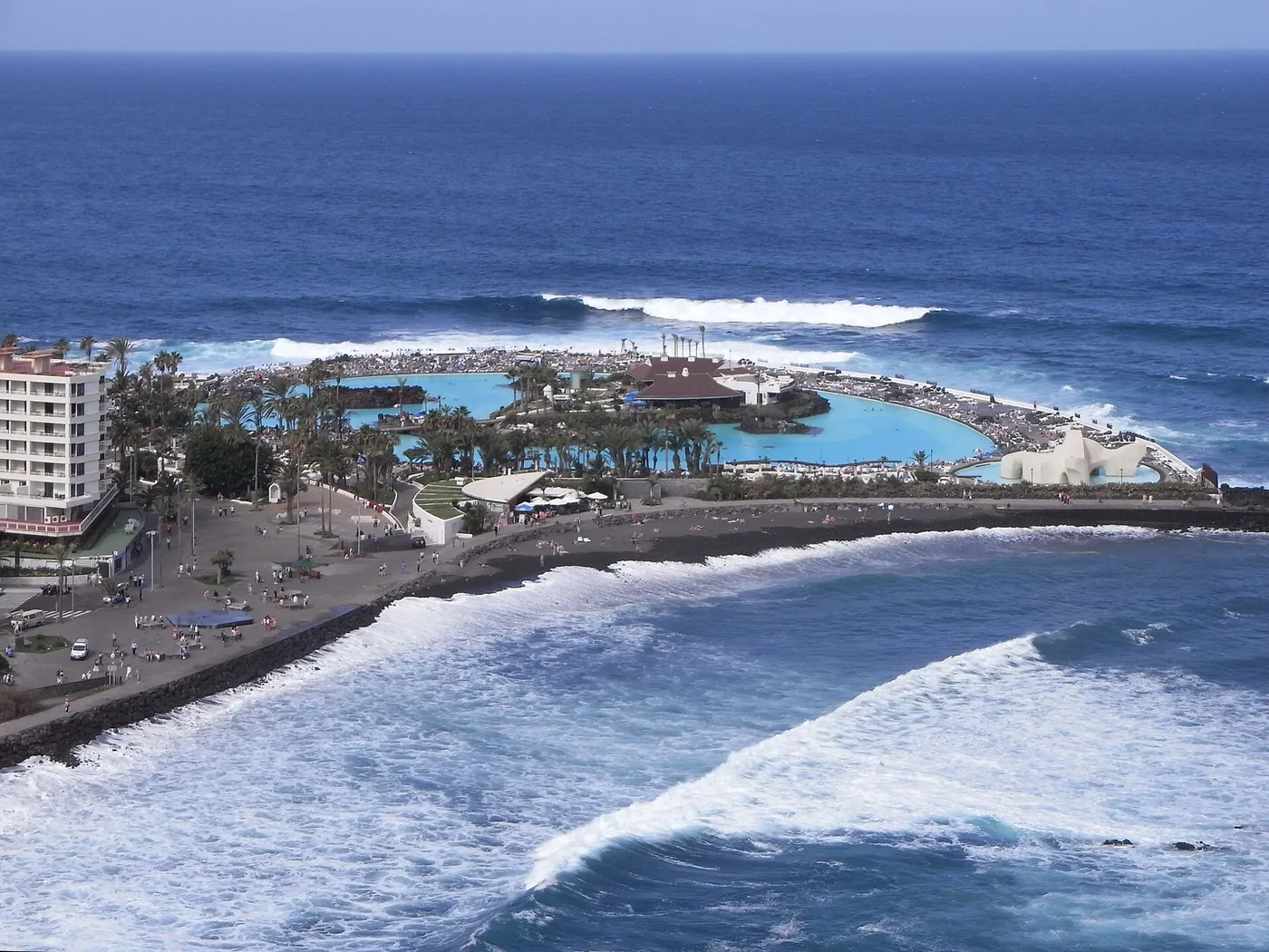 El Lago Martiánez con sus piscinas de agua turquesa y, en primer plano, la Playa Martiánez de arena oscura con olas rompiendo