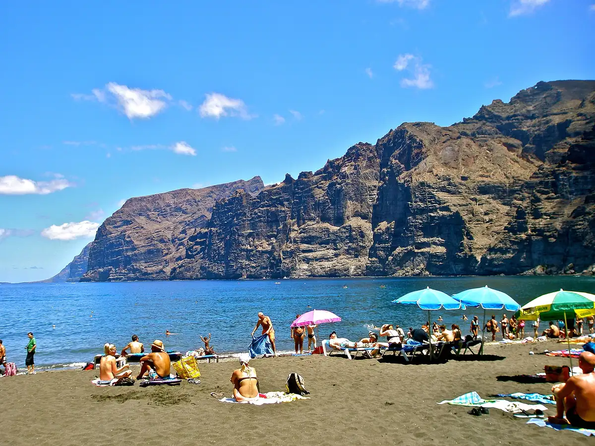 Bañistas, sombrillas y hamacas en la arena negra de la playa de Los Guíos con los acantilados al fondo